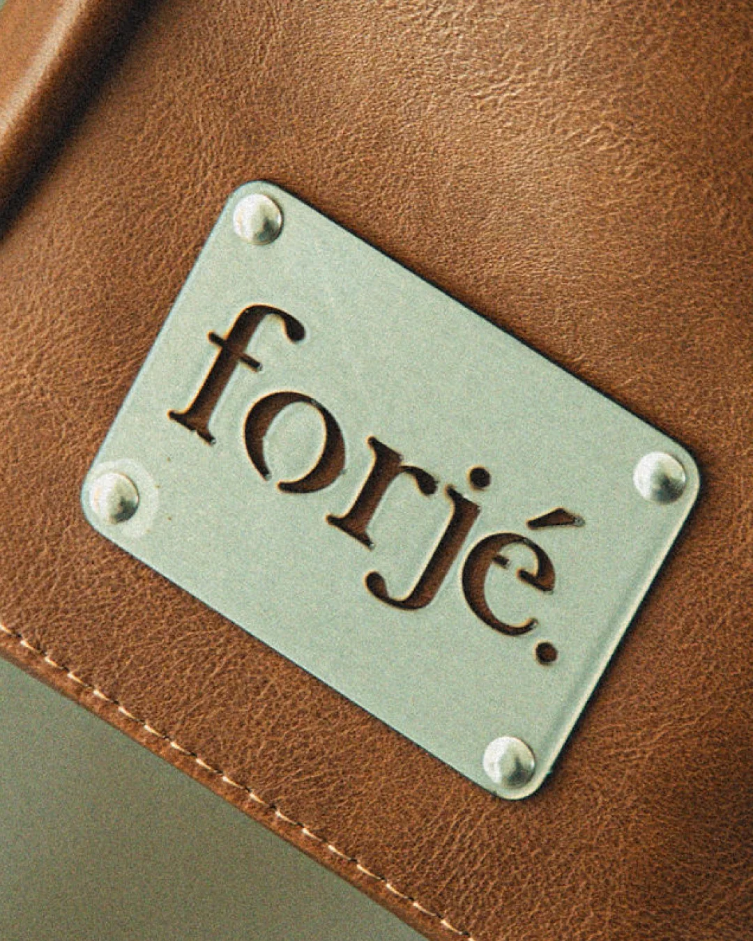 Forjé — placa metálica remachada sobre cuero, con el logotipo calado.